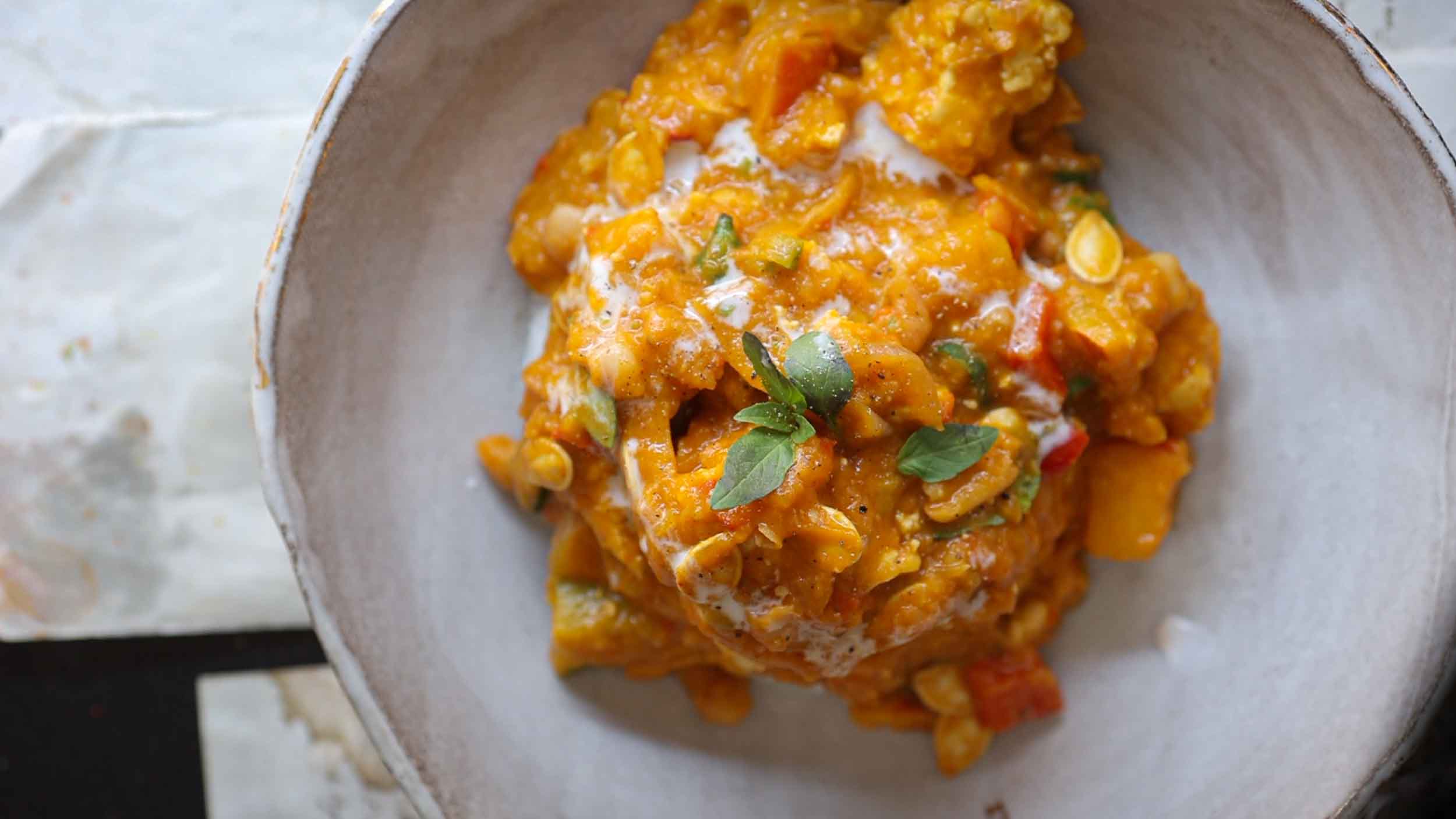 A bowl of vibrant pumpkin curry garnished with fresh green herbs. The curry has a creamy texture with visible chunks of vegetables and a drizzle of coconut milk on top, adding to its rich and aromatic appearance.